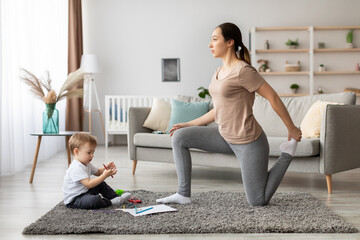In a bright and inviting living room, a mother performs a stretch while kneeling. Her young child is seated nearby, playing with colorful toys on the rug.