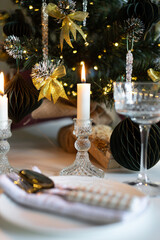 A beautifully set table for a festive event. Two lighted candles, elegant glassware and decorative ornaments are placed near the Christmas tree.