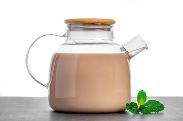 Creamy milk tea in glass teapot on table. Glass teapot filled with creamy milk tea and fresh mint leaves on table against white background.