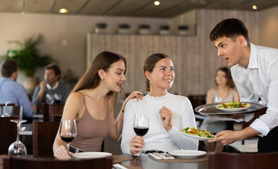 Young guy waiter brings order to couple of young women in restaurant