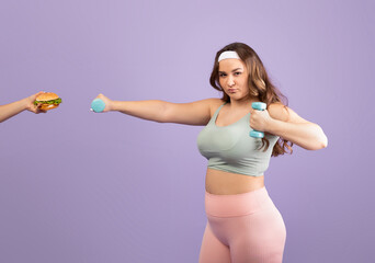 A serious plus size young woman in sportswear lifts a dumbbell in one hand while a burger is...