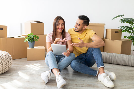A happy couple sits on the floor in their new home, sharing a moment while looking at a digital device. They are surrounded by moving boxes, suggesting they are settling in. - Powered by Adobe