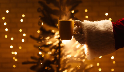 Close up of Santa Claus holding a hot drink near a Christmas tree, illuminated by warm festive...