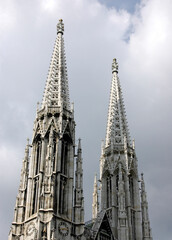 A view of the Votive Church in Vienna, Austria, showcasing its intricate Gothic architecture and towering spires against a cloudy sky.
