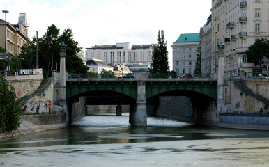 A bridge over a river in an urban setting, with buildings and trees visible. People are fishing on the side of the river.