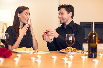 A couple enjoys a romantic dinner with pasta and wine. The man presents a surprise engagement ring, while the woman looks delighted and surprised in a cozy setting.
