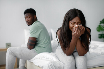 A woman sitting on the bed is crying and wiping her eyes while a man sits on the other side, looking away with a serious expression. The atmosphere is tense and filled with emotion.