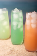refreshing trio of vibrant green, orange, and yellow drinks with ice glows against sandy beach background, evoking a chilled summer vibe with glass dew reflections, backdrop, breezy, pastel, vivid
