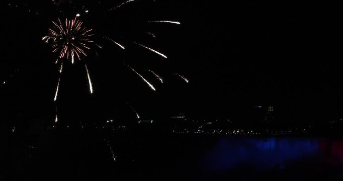Night view of Niagara Falls as fireworks burst in the sky. The waterfall is lit red and blue through the mist, with city lights in the distance. Celebration, tourism, border pride, shared wonder.