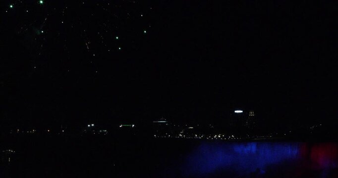 Night view of Niagara Falls as fireworks burst in the sky. The waterfall is lit red and blue through the mist, with city lights in the distance. Celebration, tourism, border pride, shared wonder.