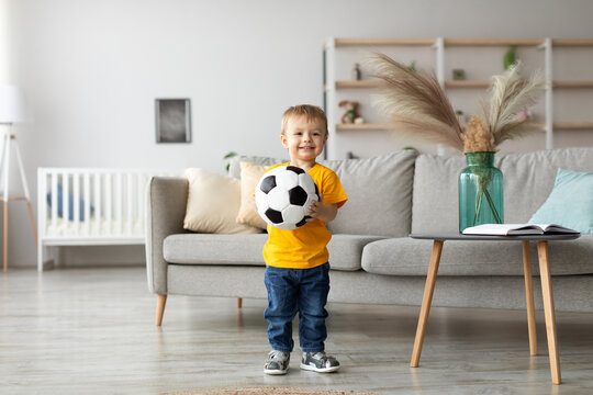 A cheerful young boy in a yellow shirt stands in a modern living room holding a soccer ball. The room features a gray couch, indoor plants, and books.