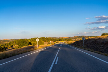 Naklejka premium Empty road winds through dry, sunlit hills under blue sky.