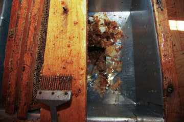 An uncapping fork rests on a honey-coated wooden beehive frame next to a stainless steel container...