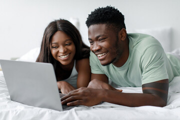 Obraz premium A man and woman share a joyful moment as they lie on a bed, smiling and engaged with a laptop. The room is bright and comfortable, creating a relaxed atmosphere for their time together.