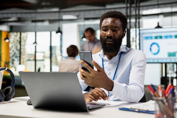Fototapeta premium African american project manager sitting with cellphone in hand, discussing with client on video call. Black male employee in modern office, taking short break to speak with family on mobile device.
