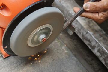 A man is sharpening iron using an orange grinding machine. Sparks appear to emerge from the...