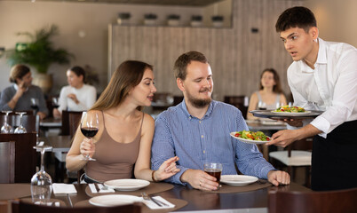 Young guy waiter brings order from couple young man and young woman in restaurant