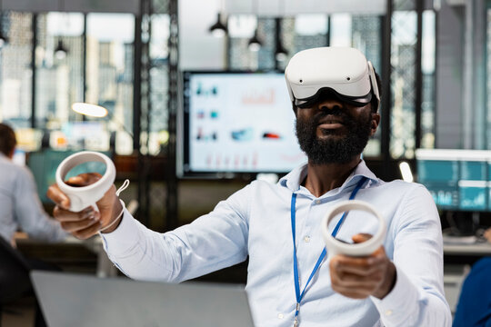Male corporate employee using VR headset and wireless controllers, interacting with 3D virtual data and numbers. Black man working in modern high tech IT company with futuristic digital technologies.