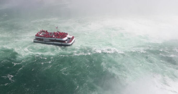 A Canadian tour boat packed with visitors in red ponchos pulls away from the base of Horseshoe Falls, surrounded by violent teal water and thick white mist. Human thrill against overwhelming power.