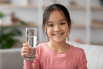 A cheerful young girl is seated in a cozy living room, holding up a glass of water. She has a warm...