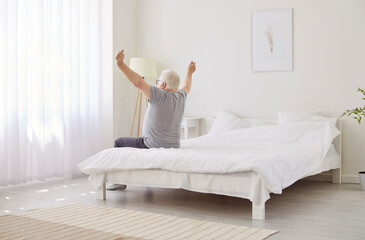 Elderly man is seen stretching comfortably in sunlit, minimalist bedroom after good nights sleep....