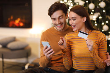 A young couple sits together by the fireplace, happily shopping online. The woman holds a credit card and points excitedly at her phone, while the man smiles at the screen.