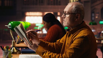 Senior male student reading a book in a study area at a public library, reviewing papers and looking for thesis citations sources. Aged man determined to graduate college.