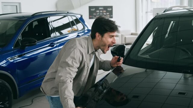 A young man stands smiling in an auto salon, holding car keys while evaluating vehicles. Behind him, SUVs are on display, showcasing the latest models.