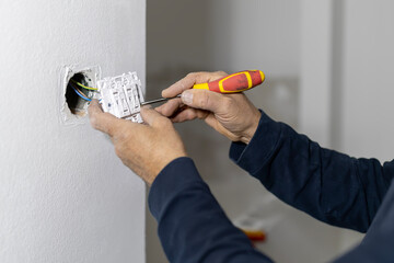 Adult man performing electrical installation work, connecting wires to a new light switch on a white wall using a screwdriver during a house renovation project