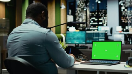 Black male employee at office uses green screen on laptop after hours, scheduling new meetings and planning the weekly agenda. Business management tasks for a company project. Camera B.
