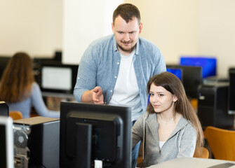 In classroom during computer courses, male teacher helps girl student complete difficult task, looks at monitor screen and corrects work. Mentor comments on students work, gives hints during task.