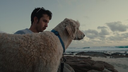 Man enjoys quiet moment with dog at the beach during sunset with gentle waves in the background