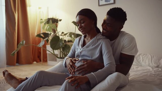 A young African American couple sits comfortably on a bed, sharing a joyful moment. The husband gently holds his wife's pregnant belly, creating a tender atmosphere in their home.
