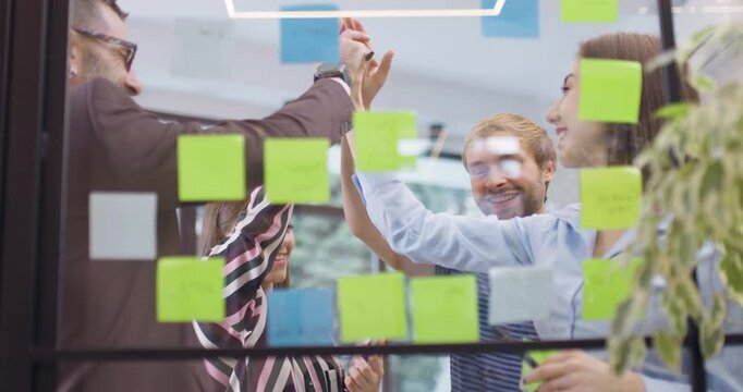 Work colleagues discussing startup project ideas and giving high five. Team of caucasian business people at glass wall with sticky notes. Scrum with colorful stickers. Low angle view.