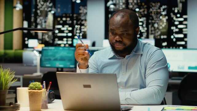 Black general manager envisioning a tactical solution and playing with pen, working late alone in office. Guy engaging in critical thinking, evaluating options and solutions for dilemma. Camera B.
