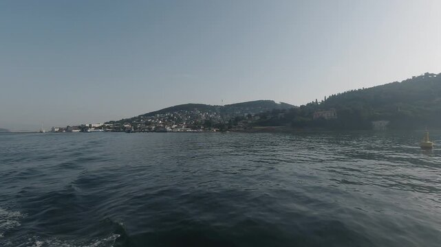 Boat-level tracking shot across shimmering water toward Adalar (Princes&rsquo; Islands). Backlit sun, soft haze, and quiet waves reveal a hillside town and pier, evoking summer calm, distance, and approach.
