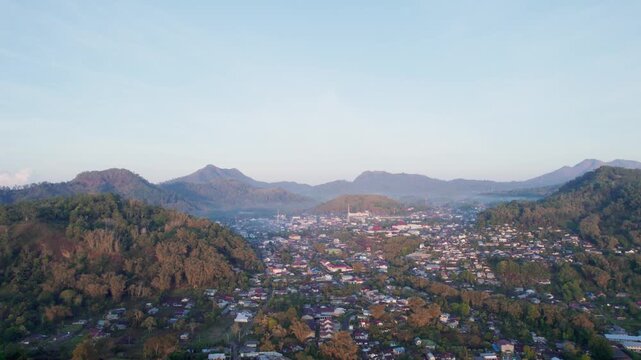 High aerial drone footage of the town of Bajawa, Flores, Indonesia. The town is nestled in a large valley surrounded by mountains. A light mist hangs over the landscape at golden hour sunrise.