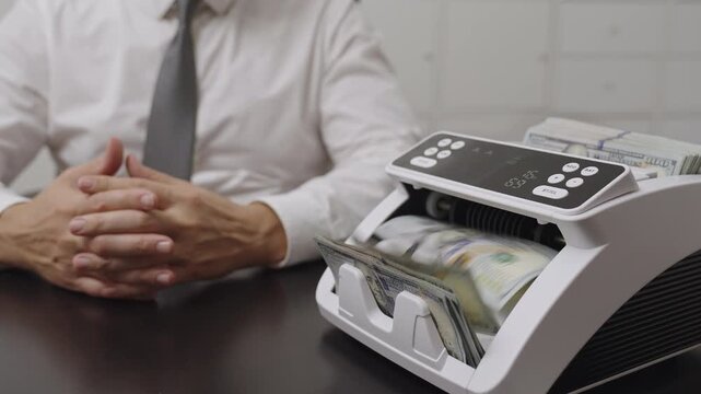 A businessman in a white shirt and tie counts U.S. dollar bills using an automatic money counting machine on a dark office desk. Concept of finance, banking, investment, and wealth management.