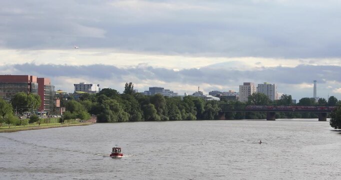 Wide view along Frankfurt&rsquo;s Main River: a small boat heads upstream, a lone rower glides opposite, a red commuter train crosses the bridge, and a plane cuts the layered sky&mdash;everyday motion and calm.