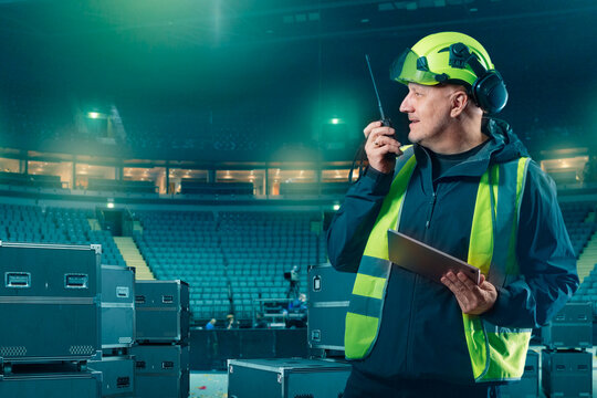 Stage supervisor in safety gear communicates via walkie-talkie while holding tablet during concert equipment setup in arena. Stage Equipment Installation. - Powered by Adobe