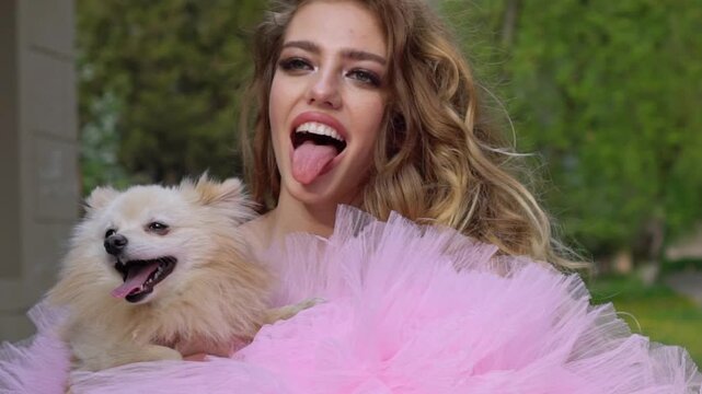 Young woman in a pink tulle dress holding a fluffy dog outdoors. Playful fashion portrait with happiness and friendship. Funny beauty girl show tongue. Fun happy emotions.
