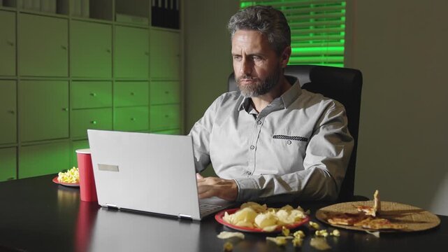 Man working on laptop late at night. Snacks and pizza on the desk. Focused on computer in dark office. Evening work with casual setup. Late-night productivity and dedication.
