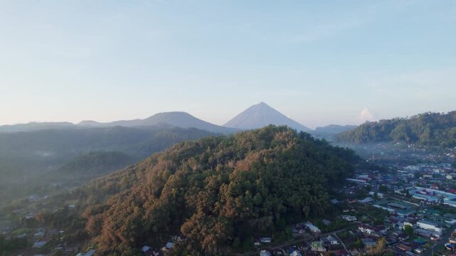 Aerial drone footage flying over a forest hill, revealing the village of Bajawa, Flores. The iconic Mount Inerie volcano stands in the background, illuminated by beautiful golden hour sunrise light.