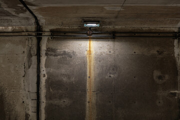 Gritty concrete wall of underpass features long vertical rust streak beneath single small overhead fluorescent light, showing horizontal pipes. Evokes urban decay, and moody utilitarian ambiance.
