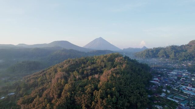 Aerial drone footage flying over a forest hill, revealing the village of Bajawa, Flores. The iconic Mount Inerie volcano stands in the background, illuminated by beautiful golden hour sunrise light.
