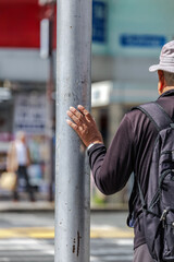 Older man with backpack waits at crossing with eyes fixed on opposite side and one hand resting on metal streetlight pole. Scene depicts moment pause under bright daylight sun amid busy urban streets.