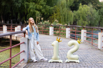 Smiling girl leaning on railing beside number twelve birthday balloons on a park bridge