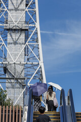 Three people escalator headed for tall tower with lattice framework set on clear blue sky. Casual dress and sun umbrellas capture an everyday moment.