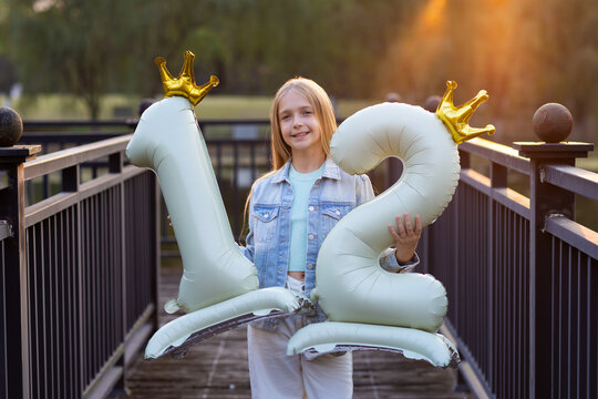 Smiling girl holding number twelve balloons with crowns on a bridge at sunset