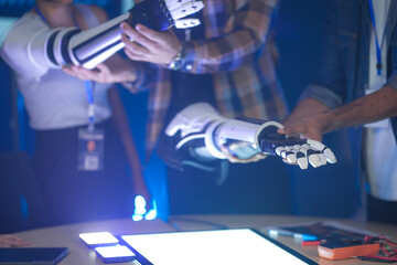 An Indian woman wearing a robotic arm prototype shakes hands with another robotic arm held by a colleague. A diverse tech team smiles, collaborating in a lab.